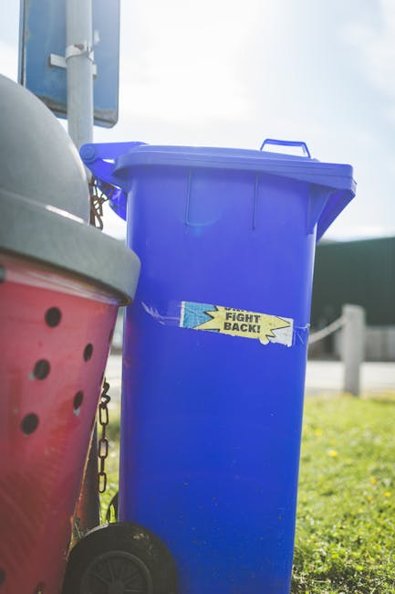 A large blue wheeled rubbish bin made of durable plastic with a closed lid, situated outdoors on a grassy area, with a yellow and blue sticker that reads 'FIGHT BACK!' affixed to its front. To the lef